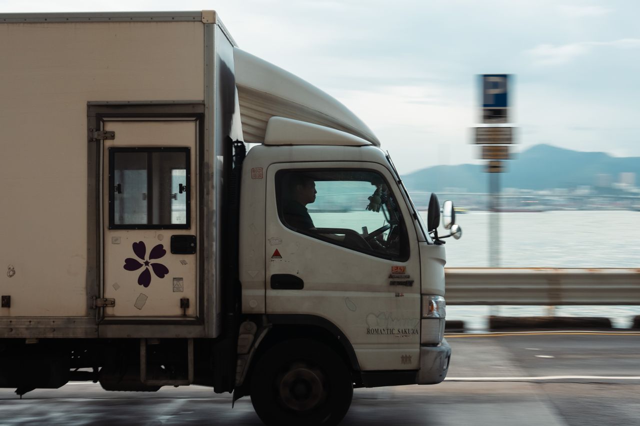 hero-img-01 A delivery truck driving along a coastal road with a mountain backdrop on a clear day.