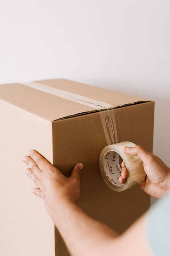 services-01 Crop anonymous male using tape to seal packed cardboard box against white wall while moving into new place