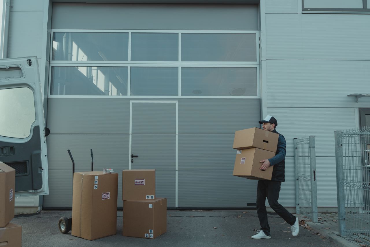 gallery-1 A delivery person carrying cardboard boxes outside a warehouse facility.
