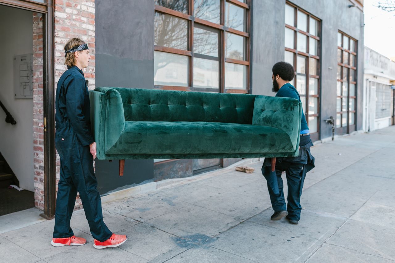 services-03 Two men carrying a green sofa on a city street, showcasing teamwork and moving services.